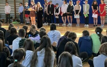 Na zdęciu widzimy grupę uczni&oacute;w na sali gimnastycznej podczas apelu z okazji 20 rocznicy wejścia Polski do UE. W tle dekoracja na niebieskim tle , biało-czerwona flaga, Białe gołębie , obok flaga UE i złote gwiazdy w kształcie koła