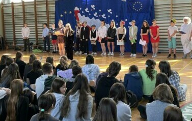 Na zdęciu widzimy grupę uczni&oacute;w na sali gimnastycznej podczas apelu z okazji 20 rocznicy wejścia Polski do UE. W tle dekoracja na niebieskim tle , biało-czerwona flaga, Białe gołębie , obok flaga UE i złote gwiazdy w kształcie koła