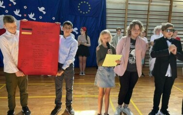 Na zdęciu widzimy grupę uczni&oacute;w na sali gimnastycznej podczas apelu z okazji 20 rocznicy wejścia Polski do UE. W tle dekoracja na niebieskim tle , biało-czerwona flaga, Białe gołębie , obok flaga UE i złote gwiazdy w kształcie koła