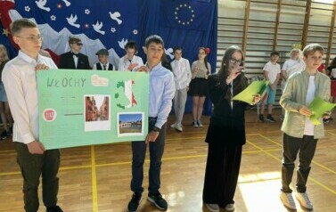 Na zdęciu widzimy grupę uczni&oacute;w na sali gimnastycznej podczas apelu z okazji 20 rocznicy wejścia Polski do UE. W tle dekoracja na niebieskim tle , biało-czerwona flaga, Białe gołębie , obok flaga UE i złote gwiazdy w kształcie koła