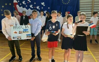 Na zdęciu widzimy grupę uczni&oacute;w na sali gimnastycznej podczas apelu z okazji 20 rocznicy wejścia Polski do UE. W tle dekoracja na niebieskim tle , biało-czerwona flaga, Białe gołębie , obok flaga UE i złote gwiazdy w kształcie koła