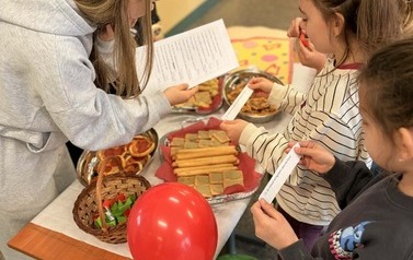 Na zdjęciu widzimy uczni&oacute;w na holu szkolnym podczas przerwy. Uczniowie biorą udział w quizie wiedzy na temat Włoch. Dookoła znajduje się dekoracja w kolorach włoskich, uczniowie biorą r&oacute;wnież udział w poczęstunku włoskim