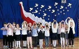 Na zdjęciu widzimy grupę uczni&oacute;w w strojach galowych za nimi na niebieskim tle biało-czerwona dekoracja. Uczniowie znajdują się na sali gimnastycznej na kt&oacute;rej odbywa się koncert pieśni patriotycznych.