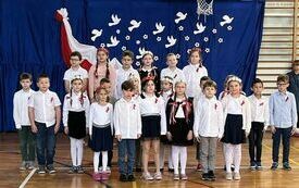Na zdjęciu widzimy grupę uczni&oacute;w w strojach galowych za nimi na niebieskim tle biało-czerwona dekoracja. Uczniowie znajdują się na sali gimnastycznej na kt&oacute;rej odbywa się koncert pieśni patriotycznych.
