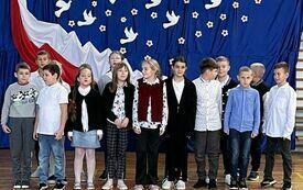 Na zdjęciu widzimy grupę uczni&oacute;w w strojach galowych za nimi na niebieskim tle biało-czerwona dekoracja. Uczniowie znajdują się na sali gimnastycznej na kt&oacute;rej odbywa się koncert pieśni patriotycznych.