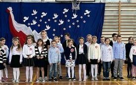 Na zdjęciu widzimy grupę uczni&oacute;w w strojach galowych za nimi na niebieskim tle biało-czerwona dekoracja. Uczniowie znajdują się na sali gimnastycznej na kt&oacute;rej odbywa się koncert pieśni patriotycznych.