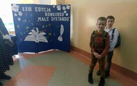 Na zdjęciu widzimy dw&oacute;ch uczni&oacute;w na korytarzu szkolnym za nimi w tle dekoracja z napisem Małe dyktando.
