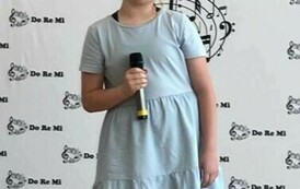 Na zdjęciu widzimy  uczennicę trzymającą w ręku mikrofon,  śpiew piosenkę na konkursie, za nią w tek dekoracja z napisami do re mi