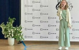 Na zdjęciu widzimy  uczennicę trzymającą w ręku mikrofon,  śpiew piosenkę na konkursie, za nią w tek dekoracja z napisami do re mi