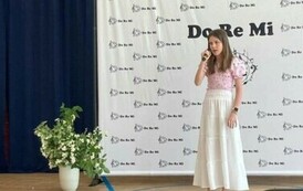 Na zdjęciu widzimy  uczennicę trzymającą w ręku mikrofon,  śpiew piosenkę na konkursie, za nią w tek dekoracja z napisami do re mi