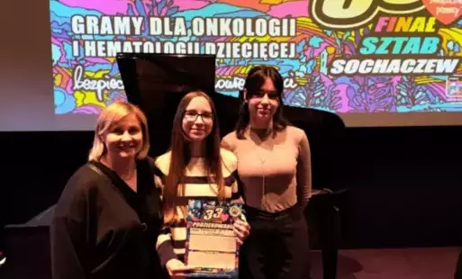 Na zdjęciu widzimy nauczyciela i dwie uczennice, które pozują do zdjęcia za nimi w tle ścianka z 33 finałem WOŚP