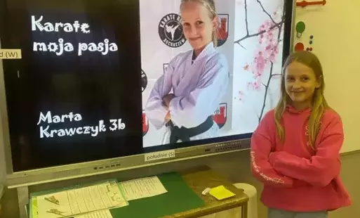 Na zdjęciu widzimy uczennicę pozującą do zdjęcia za nią w tle tablica interaktywna z napisem Karate moja pasja i zdjęciem uczennicy