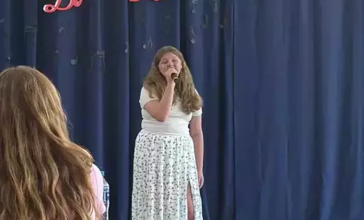Na zdjęciu widzimy uczennicę w jasnej sukience ma długie włosy w ręku trzyma mikrofon i śpiewa za nią na granatowym tle napis czerwony Do RE Mi 