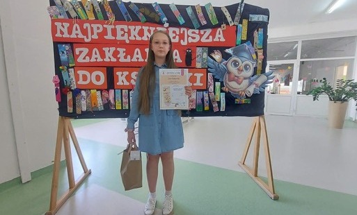 Na zdjęciu widzimy uczennice z dyplomem w ręku za nią w tle napis "Najpiękniejsza zakładka do książki".