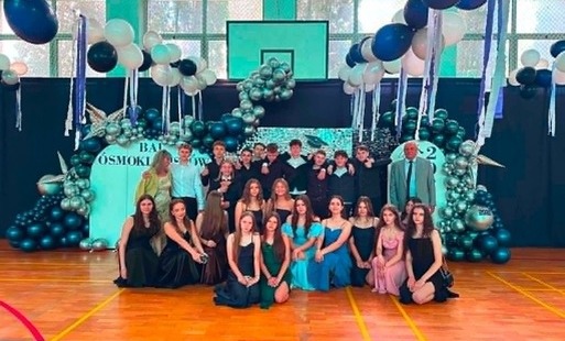 Uczniowie ósmych klas pozują do zdjęcie podczas balu, za nimi w tle ścianka dekoracyjna.