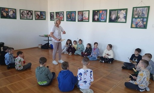 Na zdjęciu widzimy nauczyciela prowadzącego zajęcia muzyczne , wokół niego uczniowie klasy trzeciej , na scianach wiszą kolorowe obrazy.